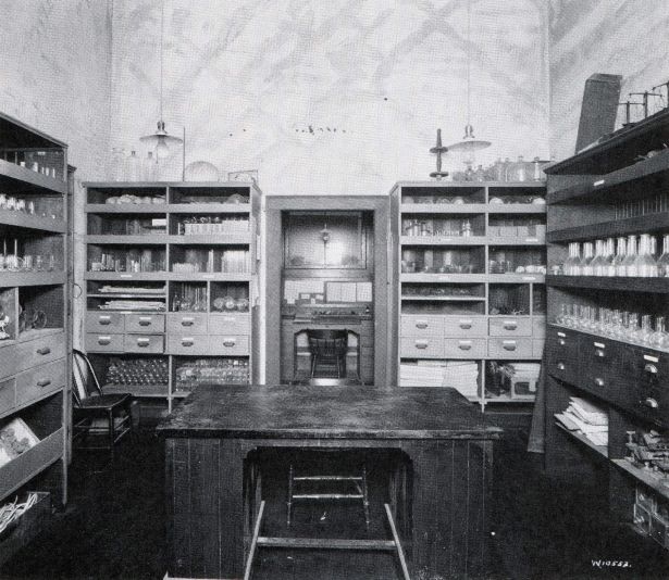 a room with chemistry equipment in shelving units along the walls and large and small desks in the middle.