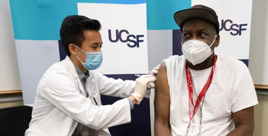 Aludino in a white coat administers a vaccine to a man in a baseball cap