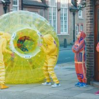 Four people in yellow, orange, and purple hooded sweatsuits acting as proteins. One person wearing sweatsuit inside large yellow bubble acting as a cell nucleus.