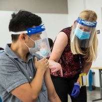 Clinard, wearing a face mask and face shield, points to a student's arm as another holds a needle.