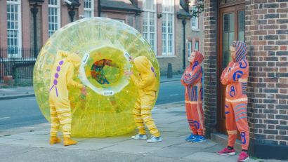 Four people in yellow, orange, and purple hooded sweatsuits acting as proteins. One person wearing sweatsuit inside large yellow bubble acting as a cell nucleus.