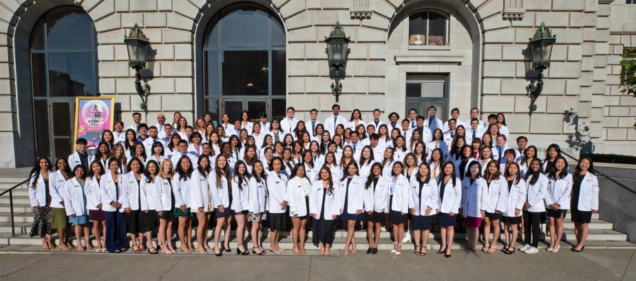 Group of approximately 122 students in white coats on the steps of a building.