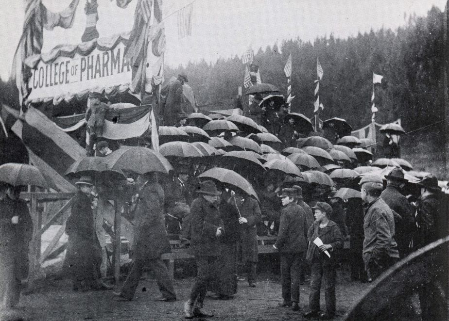 people with umbrellas gather on bleachers with a bunting-decorated sign saying College of Pharmacy.