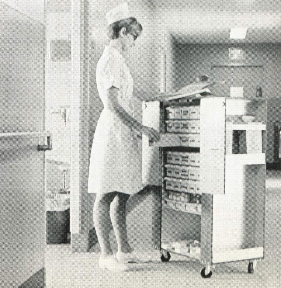 a woman in a white uniform stands next to a mobile cart with medications and consults a document.
