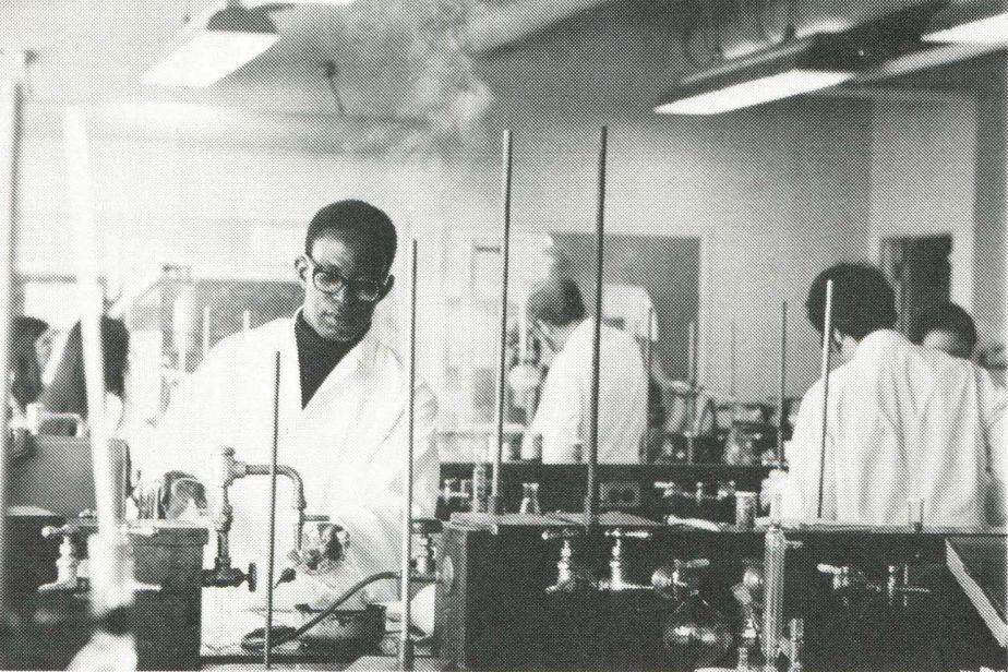 student wearing a white coat and protective eyewear works in a lab amongst other students.